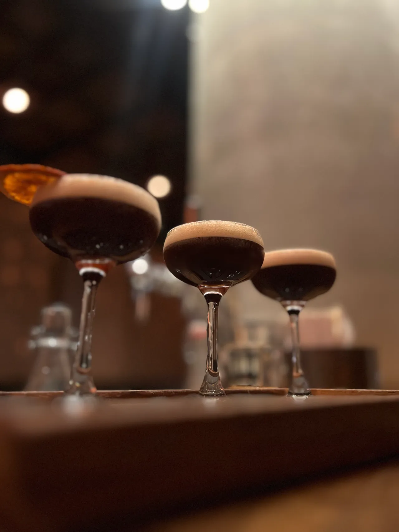 Three espresso martinis lined up on a bar rail with warm background bokeh