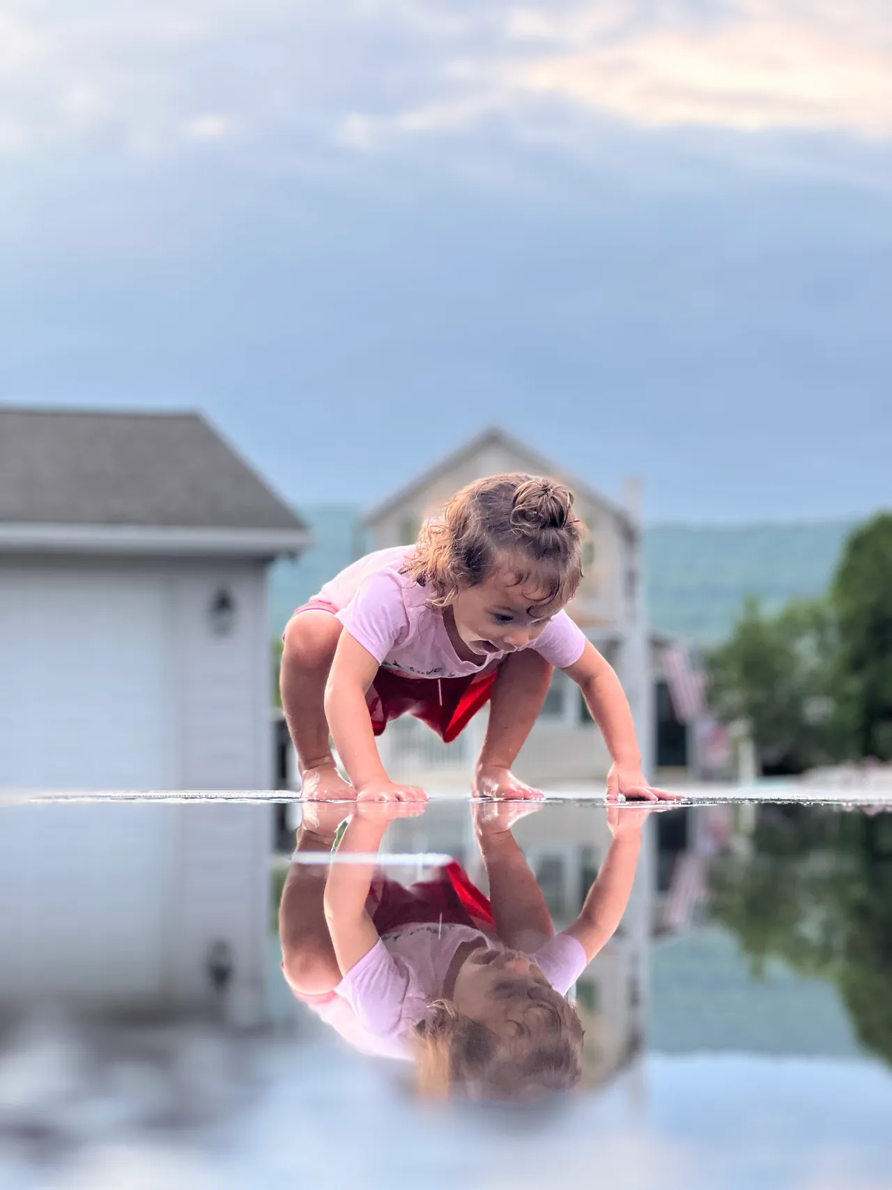 Toddler crouching at the edge of a mirror-still puddle, houses and hills reflected